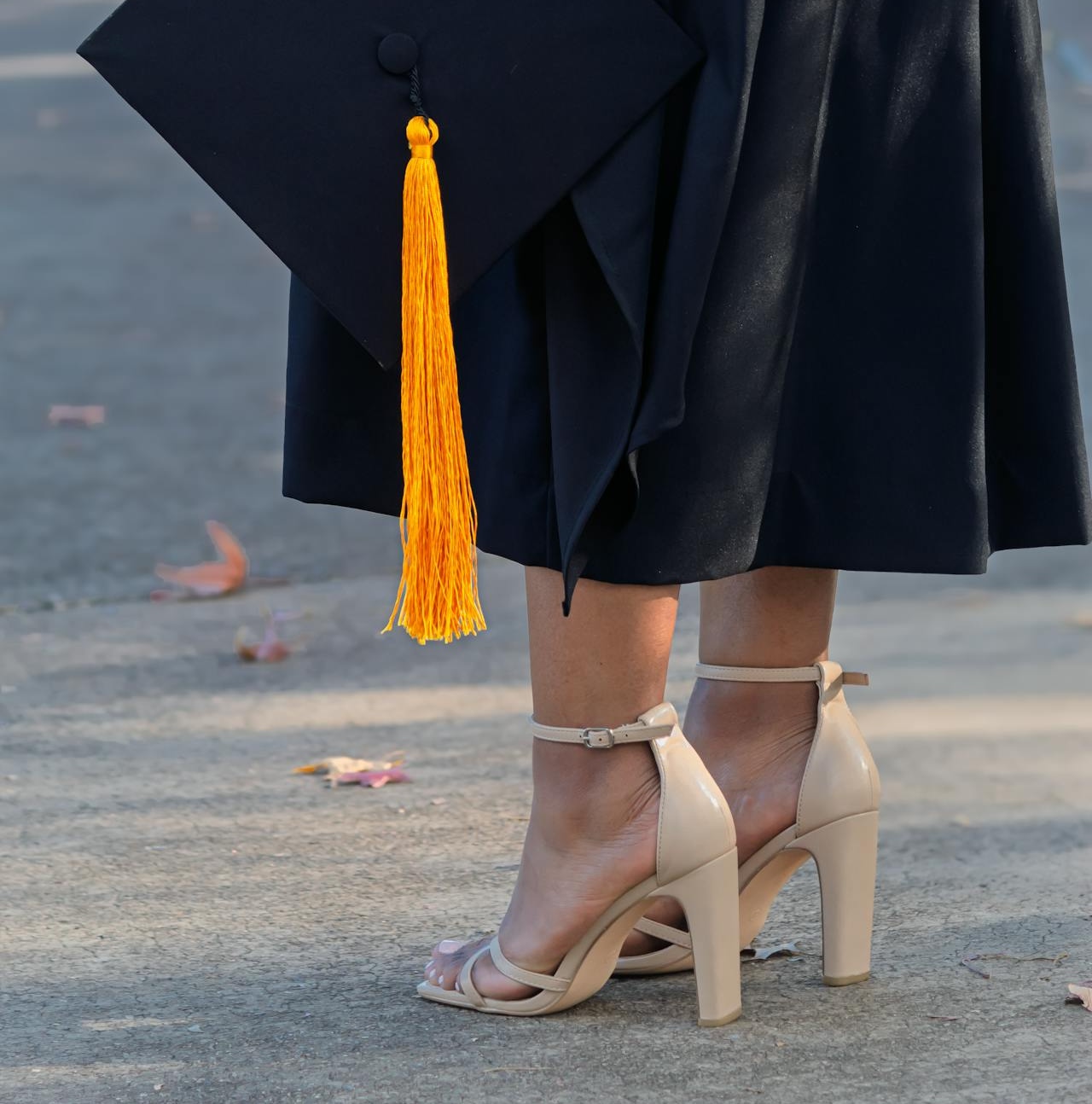 Close-up of nude block heel ankle strap sandals worn under a graduation gown with mortarboard cap and gold tassel